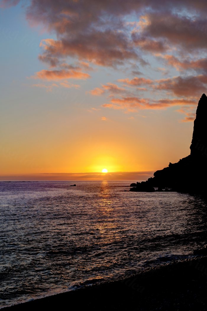 Sole che tramonta sul mare con nuvole arancioni e uno sperone di roccia scura in controluce